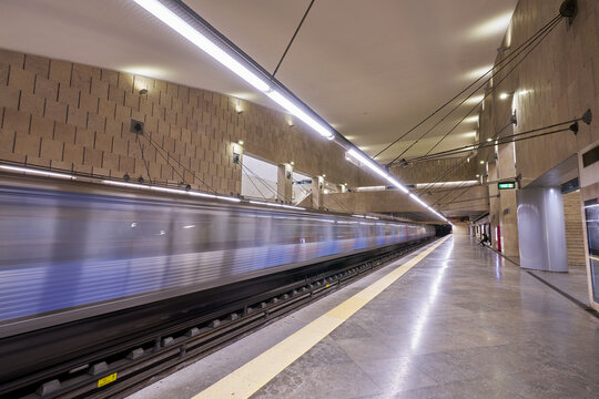 View of a motion-blurred subway train arriving at a modern underground metro station with stone-tiled walls and bright linear lighting in Lisbon, Lisbon, Portugal.