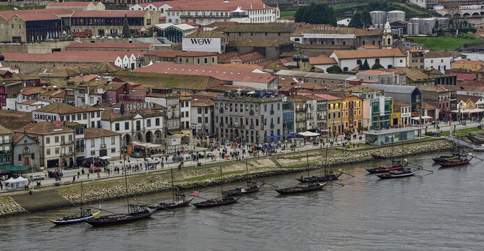 View of the historic Ribeira district along the Douro river with traditional Rabelo boats moored at the quay and people walking on the promenade in Porto, Porto District, Portugal.