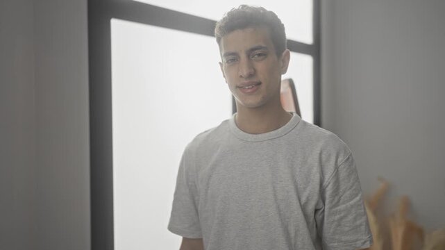 Young hispanic man smiles at camera, face visible and bare, wearing gray tshirt and standing by large window in studio; friendly ease.