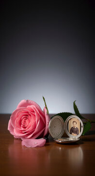 Pink rose flower beside open vintage silver locket with portrait on reflective wooden surface in dark room
