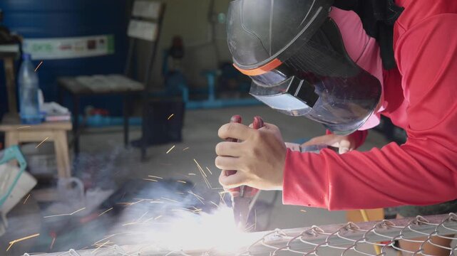 Welder wearing a protective face shield and gloves welds a steel rebar grid on the ground, with sparks and workshop tools visible in the background. Represents construction, metalwork, and industrial	