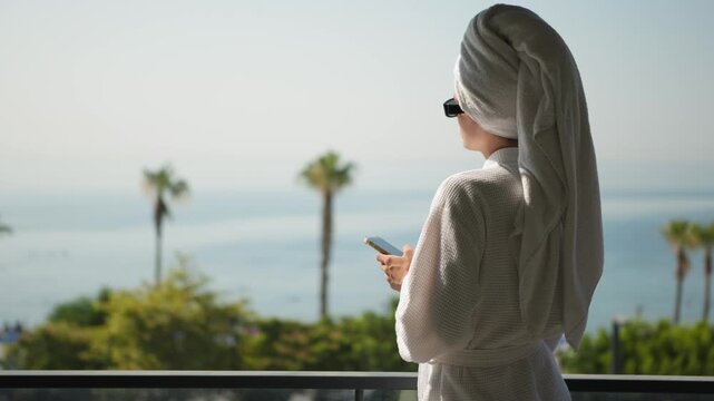 Woman in Bathrobe Messaging on Smartphone with Ocean View at Sunrise. Woman Using Smartphone in Hotel Room with Coastal View and Palm Trees at Sunrise
