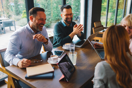 Colleagues laughing during a casual business meeting at a cafe with laptops and coffee
