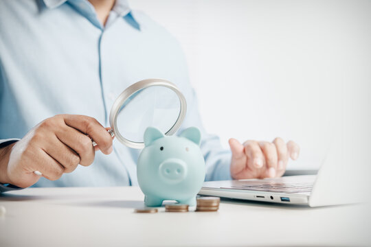 Financial planning and savings concept with magnifying glass examining a piggy bank, coins and laptop on a clean white desk, representing personal finance, budgeting and wealth management