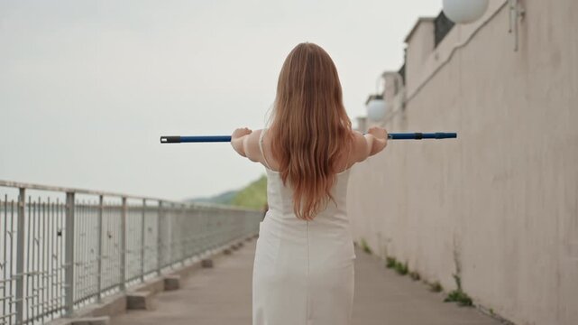 Woman in white dress walks along outdoor pathway holding blue handle of what appears to be cleaning equipment behind her back. scene features soft overcast lighting and includes elements