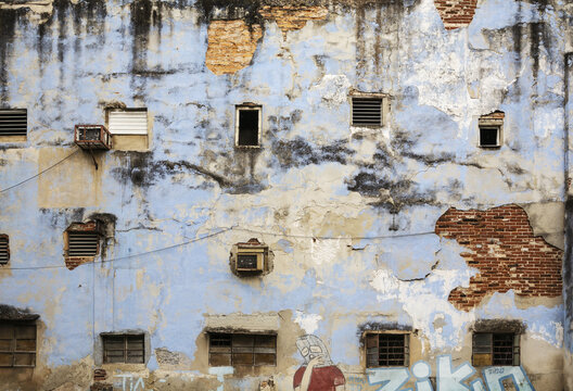 View of a weathered blue wall with peeling paint, exposed red brick, various windows, air conditioning units, and graffiti in Havana, Havana, Cuba.