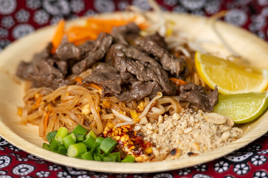 Beef Pad Thai, a popular Thai stir-fried rice noodle dish, garnished with sliced ​​beef, lime, crushed peanuts, and scallions, served in a bamboo dish