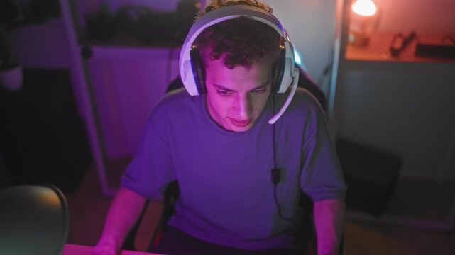 Young hispanic man wearing headset eats burger while leaning forward at a computer in studio; concentration gaming snack.