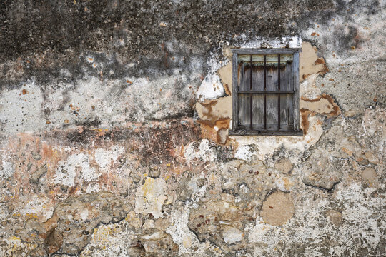 View of a weathered, textured wall with peeling paint and a small, rustic wooden window with iron bars in Gibara, Holguin, Cuba.
