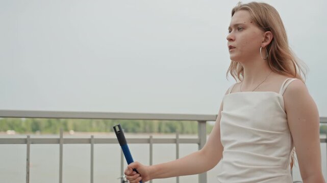 Young woman stands on bridge holding and looking at object in her hand. Woman wears white dress with thin straps against backdrop of water and distant greenery under overcast skies. scene