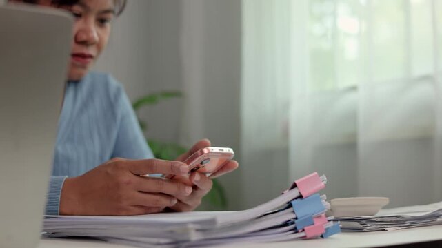 Asian female employee typing on mobile phone screen sitting at work desk beside stack of documents checking online updates and client messages during office work shift hours