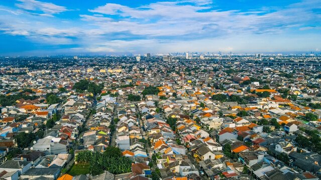 Aerial view of dense residential neighborhood with numerous rooftops, scattered trees, and a distant urban skyline under a clear blue sky in Bekasi Barat, West Java, Indonesia.