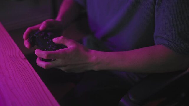 Young man holds game controller with both hands, thumbs pressing buttons on controller beside wooden desk at studio; concentration intensity.
