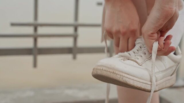 Woman ties laces of white sneaker focusing on hands and shoe details. action is captured in close up with soft lighting emphasizing texture detail as woman secures her footwear outdoors