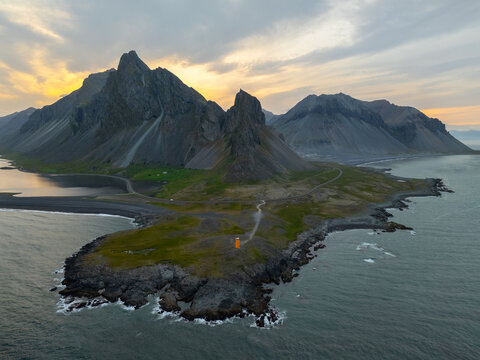 Aerial view of Hvalnes Lighthouse standing on a rocky peninsula with dramatic mountains in the background under a golden sunset sky in Sveitarfelagid Hornaf, Iceland.