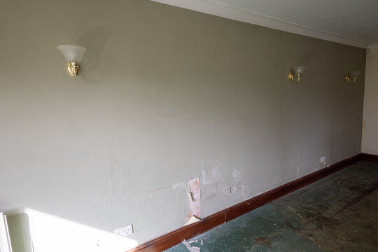 Neglected empty room with damp damaged wall peeling paint exposed plaster dirty green carpet wooden skirting and wall lights showing an older residential property needing renovation