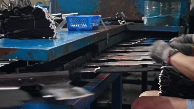 Workers seated in row fit knitted protective gloves onto metal forms moving along conveyor. Synchronized teamwork keeps industrial glove production flowing at steady pace.