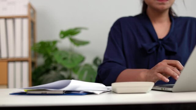 Asian female entrepreneur writing corporate strategy plan in journal book and checking electronic summary on laptop screen at work desk in quiet modern office workplace space