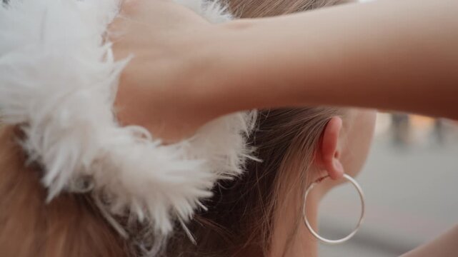Woman with long brown hair styles her loose strands into low ponytail using white fluffy scrunchie. Woman wears large hoop earrings and background is softly blurred to emphasize close
