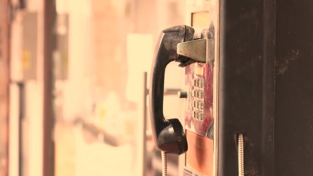 Old public payphone handset hangs on a worn metal booth along a city street