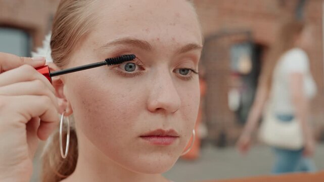 Young woman applies makeup outdoors focusing on her eye area with mascara wand. Woman wears large hoop earrings and has light colored hair tied back against urban background of brick