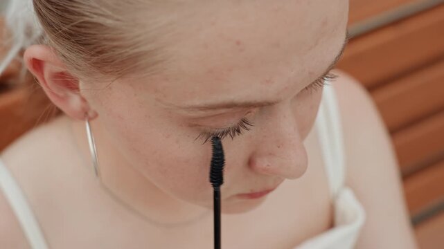 Young woman applies black mascara to her eyelashes in close up view focusing on precise movement of applying makeup. Woman wears hoop earrings and white top with wooden slats visible