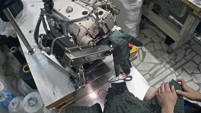 Top view closeup of worker guiding knitted work glove cuff into automatic overlock stitching mechanism on textile factory line.