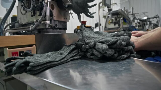 Closeup of finished knitted work gloves stacked in piles on a stainless steel workbench while a factory employee sorts pairs next to automated machinery.