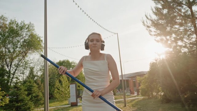 Woman in white dress and headphones dances with long blue poles outdoors on sunlit pathway surrounded by greenery. Woman moves gracefully holding props as sunlight creates warm lens flares