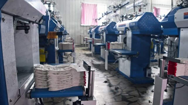 Wide shot of textile factory workshop interior with rows of automated knitting machines producing white work gloves.