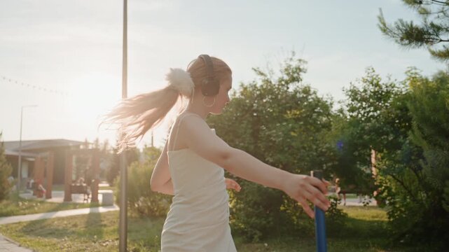 Young woman wearing headphones and white dress practices dance moves with blue pole in outdoor park setting. Woman holds long thin object while moving her arms and legs surrounded
