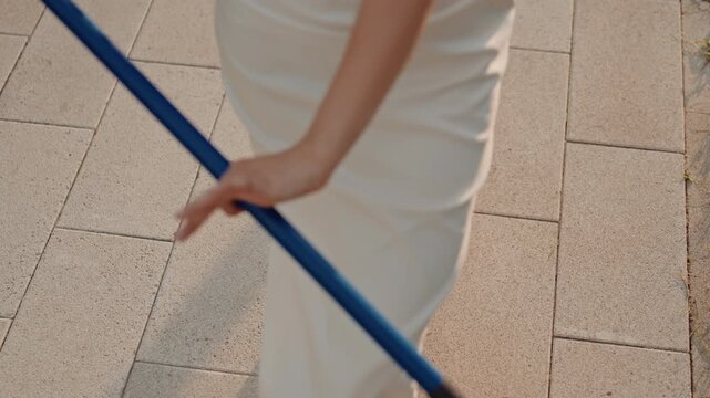 Woman in white dress sweeps ground with blue broom focusing on her lower body and feet as woman moves along paved tiles. scene emphasizes repetitive sweeping motion against outdoor