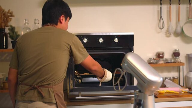 Young Man Baking Cookies at Home Kitchen, Checking Oven with Stand Mixer Nearby