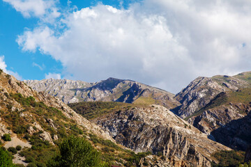 Fototapeta premium Rugged mountains stretch beneath a vibrant blue sky. Clouds drift above jagged peaks and rocky ridges. Earth tones dominate the landscape, with green accents. Sunlight highlights the textured stone