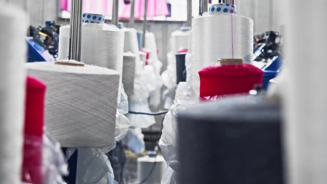 Close up of thread unwinding from large spools inside industrial clothing production facility. Textile yarn feeds continuously into manufacturing equipment.
