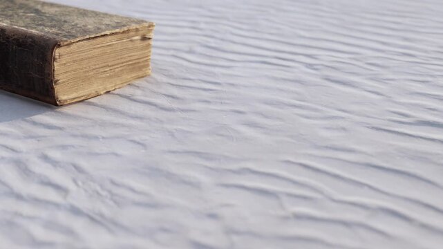 A vintage leather bound book lies quietly on soft white fabric, the intricate textures creating a serene backdrop. The warm hues of the cover contrast gently with the fabrics smoothness.