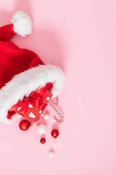 Red Santa Claus hat with Christmas gift box, candy cane and baubles on pink background. Flat lay vertical holiday composition with copy space. Creative New Year greeting card template or winter concep