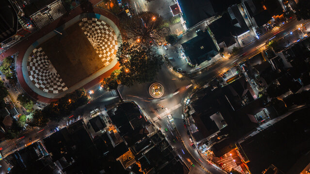 Aerial view of an illuminated city intersection at night featuring a checkered pattern plaza, surrounding buildings, and light trails from moving vehicles in Sukabumi, Indonesia.