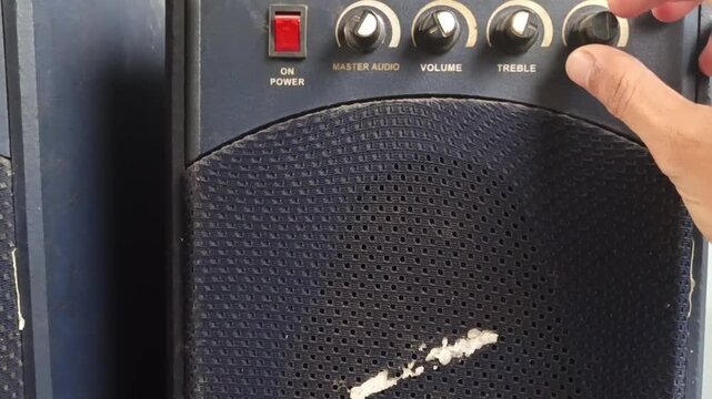 A hand is adjusting the audio controls on a black speaker amplifier, showing the volume and sound settings in the room.