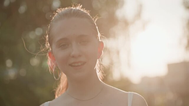 Young woman with light hair tied back wears white spaghetti strap top and silver hoop earrings standing outdoors in soft golden sunlight. Woman occasionally raises her arms or makes hand