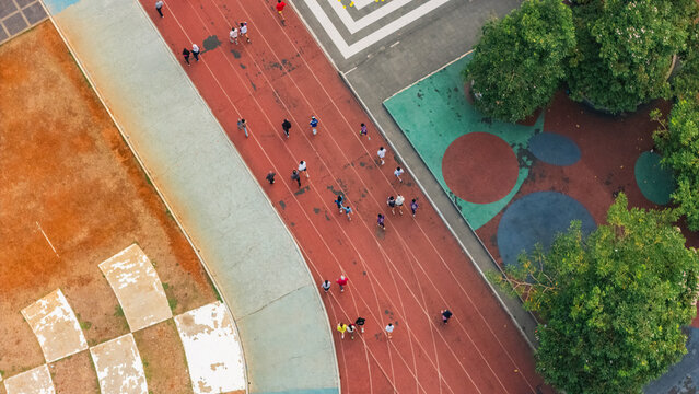 Aerial view of a running track with people exercising on the red surface, surrounded by green trees and paved areas in Indonesia.