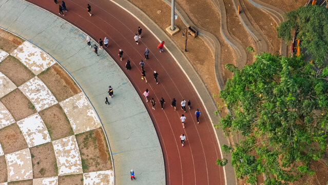Aerial view of people running on a red athletic track surrounded by lush green trees and a paved area with a checkered pattern in Indonesia.