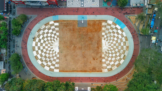 Aerial view of a public square featuring a large checkered pattern, surrounded by a running track, trees, and urban infrastructure in Indonesia.