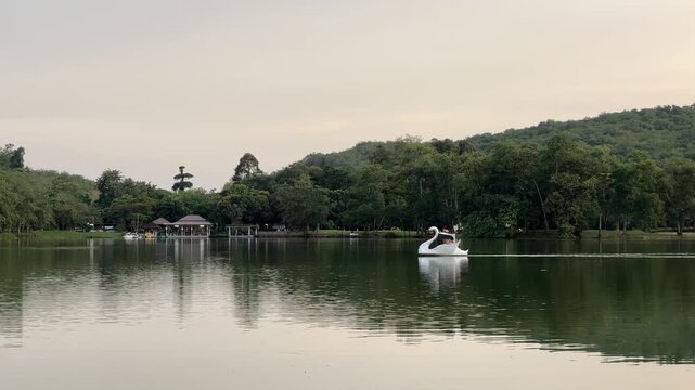 White swan pedal boat on a calm lake with forest and hill reflections at dusk.