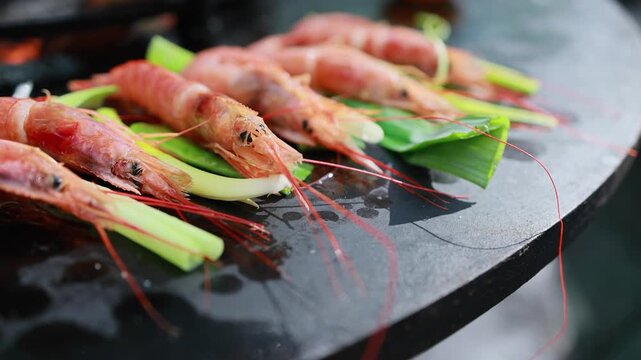 Fresh prawns with green onion cooking on hot black grill plate. Seafood barbecue preparation outdoors. Grilled shrimp with leek stems, healthy food and street food concept.