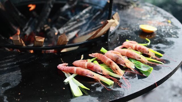 Fresh prawns with green onion cooking on hot black grill plate. Seafood barbecue preparation outdoors. Grilled shrimp with leek stems, healthy food and street food concept.