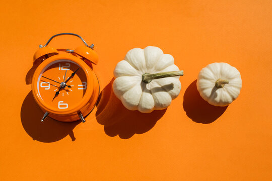 Orange alarm clock with three white decorative pumpkins on two tone beige and orange background for autumn season and daylight saving time concept