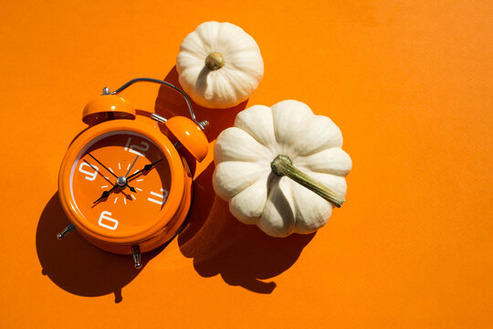Orange alarm clock with three white decorative pumpkins on two tone beige and orange background for autumn season and daylight saving time concept