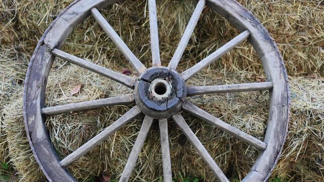 Old wooden wagon wheel leaning against hay bales on farm. Vintage rustic cart wheel with spokes. Rural countryside background, agriculture and traditional farming concept
