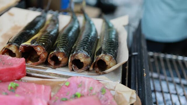Smoked mackerel fish and fresh tuna steaks on tray at street food market stall. Traditional seafood delicacy displayed on parchment paper near grill.